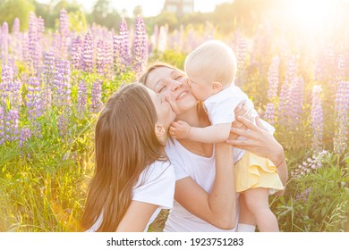 Young Mother Embracing Her Kids Outdoor. Woman Baby Child And Teenage Girl Sitting On Summer Field With Blooming Wild Flowers Green Background. Happy Family Mom And Daughters Playing On Meadow