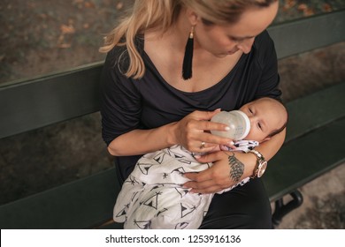 Young Mother Feeding Her Adorable Baby Boy With A Formula Bottle While Sitting Outside On A Park Bench