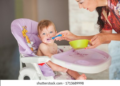 Young Mother Feeding Her Baby Girl With A Spoon. Adorable Baby Eating Food On Kitchen. Little Baby Making A Mess.