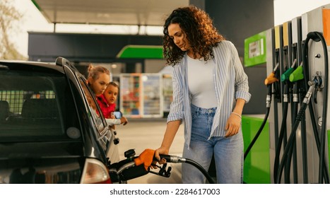 A young mother fills up gas tank at a gas station while her daughters look out the car window
