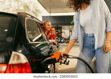 A young mother fills up gas tank at a gas station while her daughters look out the window