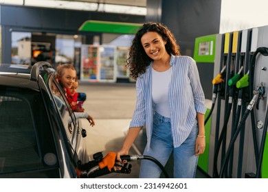 Una joven madre llena un tanque de gas en una gasolinera mientras sus hijas miran por la ventana