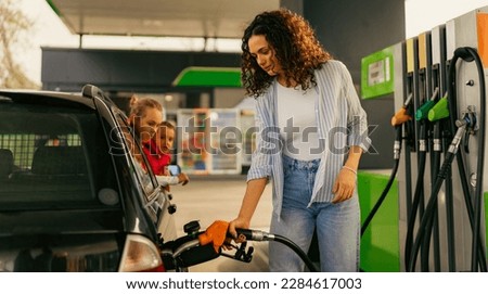 A young mother fills up gas tank at a gas station while her daughters look out the car window