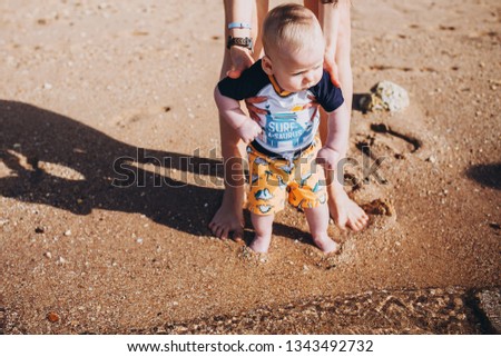 Young mother for the first time for a young boy shows sand on the beach, the sea and gives a try to touch the water