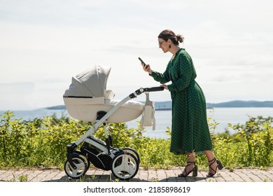 A Young Mother In A Green Dress Walks With Her Baby. Mom Takes A Picture Of A Baby In A Stroller While Walking In The City.