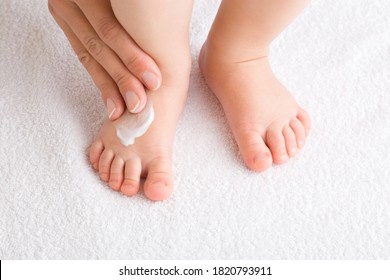 Young Mother Hand Applying White Moisturizing Cream On Baby Foot. Infant Legs Standing On White Towel. Care About Children Clean And Soft Body Skin. Closeup. Front View.