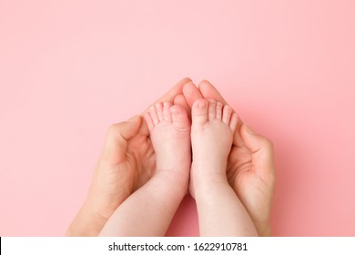 Young Mother Hands Holding Her Infant Little Feet On Pink Table Background. Pastel Color. Lovely, Emotional Moment. Closeup. Baby Legs Care.