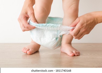 Young mother hands wearing white baby diaper pants. Little kid legs standing on floor. Closeup. Front view.
