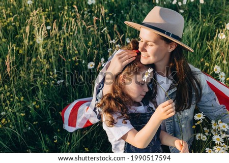 Young mother in hat and cute small daughter kid holds usa United States flag sit on green meadow at summer day. Smiling free proud independent patriotic family feeling freedom, independence concept