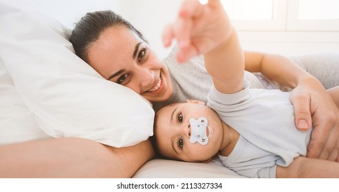 Young mother having fun taking selfie with her baby in bed at home - Soft focus on kid face - Powered by Shutterstock