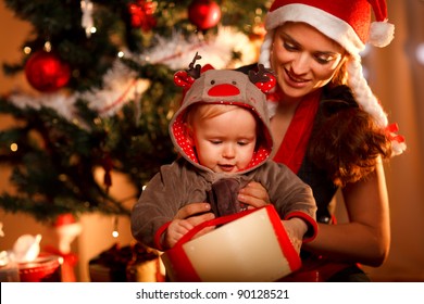 Young Mother Helping Interested Baby Open Present Box At Christmas Tree