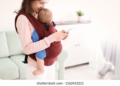 Young Mother With Her Baby In Ergo Carrier Using Mobile Phone Indoors