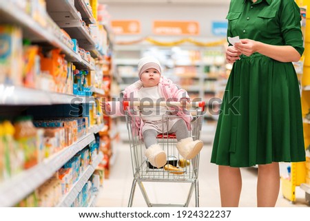 A young mother and her baby sitting in a grocery cart choose food on a supermarket shelf. Side view. Close up. The concept of shopping and parenting.
