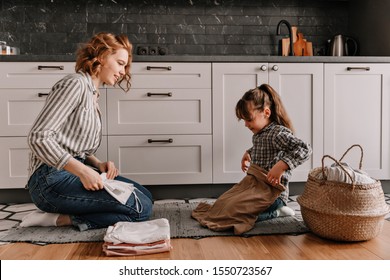 Young Mother And Her Cute Daughter Gently Fold Clothes Sitting On Kitchen Floor