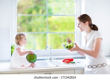 Young Mother And Her Cute Toddler Daughter Cooking Together In A Beautiful Sunny Kitchen With A Big Window