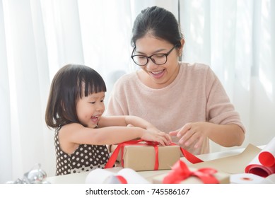 Young Mother And Her Daughter Wrapping A Gift Box For Birthday, Christmas And New Year In A White Room. Happy Asian Family At The House. Copy Space.