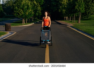 Young Mother With Her Kid In A Jogging Stroller During Jogging In A Public Park. Togetherness Mom With Son During Active Running, Happy Motherhood