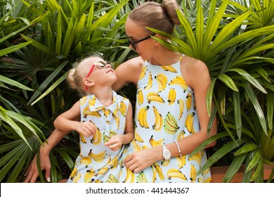 Young Mother And Her Little Daughter In Tropical Garden Among Green Foliage. Both Blonde And Wearing The Same Dress.