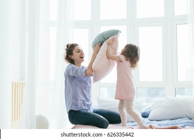 Young Mother And Her Little Daughter Playing With Cushions On Bed. Funny Pillow Fight. Soft Pastel Colors, Pink And Blue. Selective Focus. Play Together And Enjoy The Moment! Family Time On Weekend. 