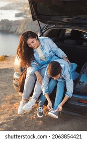 A Young Mother And Her Teenage Son In Denim Clothes Enjoy The Springtime, Sitting In The Open Trunk And Hugging. The Teenager Ties The Laces On The Sports Shoes. Mother Teaches To Tie Shoelaces. 