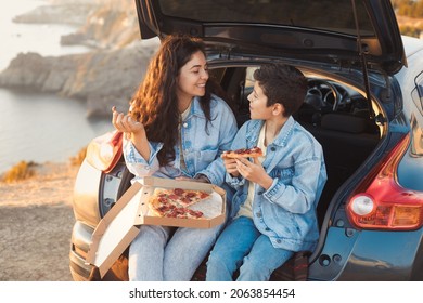 A young mother and her teenage son in denim clothes enjoy the springtime on the top of the hill, sitting in the open trunk of a car and eating pizza. Spanish tourists woman and child at vacation. 