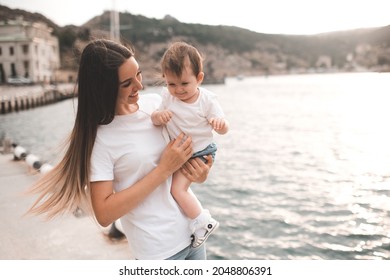 Young Mother Hold Baby Girl 1 Year Old Wear Casual Clothes Walk In City Street Over Seafront. Motherhood. Happy Woman With Child Together Outdoor. 