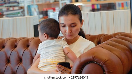 Young Mother Holding Baby Boy And Browsing On Mobile Phone Sitting On Couch