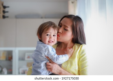 Young Mother Holding Baby In Her Arms And Kissing Him At Home. Happy Mixed Race Asian-German Woman And Child. Mother’s Day, Love Mom Concept.