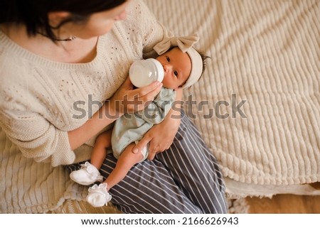 Young mother holding baby on hands in bed feeding with milk in plastic bottle top view close up. Motherhood. Newborn child with woman wake up and eating in morning at home. Healthy infant nutrition.