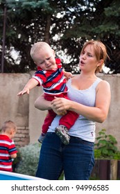 Young Mother Holding Grumpy Crying Toddler Boy Outdoors In The Garden.