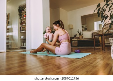 Young mother holding her adorable baby on an exercise mat. Happy mom working out with her little baby at home. New mom bonding with her baby during her post-natal fitness routine.