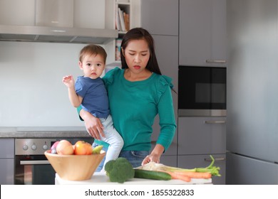 Young Mother Holding Her Baby While Cooking At Kitchen Home. Mixed Race Asian-German Family Mom And Little Son. Busy And Stressed Mommy Housewife Overwork Concept.