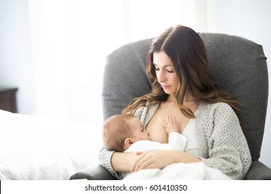 A Young Mother Holding Her Baby Child. Mom Nursing Baby. Woman And New Born Boy Relax In A White Bedroom With Rocking Chair And Blue Crib.