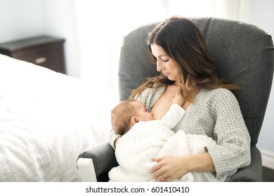 A Young Mother Holding Her Baby Child. Mom Nursing Baby. Woman And New Born Boy Relax In A White Bedroom With Rocking Chair And Blue Crib.