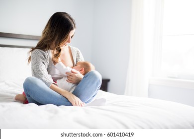 A Young mother holding her baby child. Mom nursing baby. Woman and new born boy relax in a white bedroom with rocking chair and blue crib.