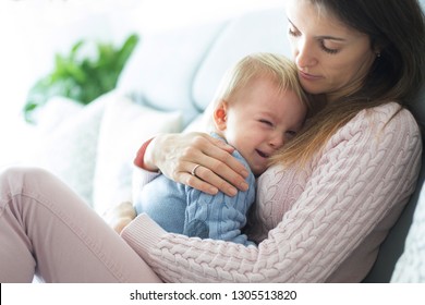 Young Mother, Holding Her Crying Toddler Boy, Hugging Him At Home, Trying To Calm Him Down In Sunny Living Room