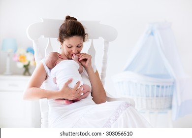 Young Mother Holding Her Newborn Child. Mom Nursing Baby. Woman And New Born Boy Relax In A White Bedroom With Rocking Chair And Blue Crib. Nursery Interior. Mother Breast Feeding Baby. Family At Home