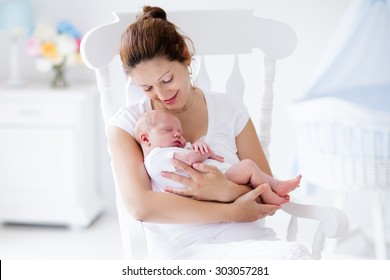 Young mother holding her newborn child. Mom nursing baby. Woman and new born boy relax in a white bedroom with rocking chair and blue crib. Nursery interior. Mother breast feeding baby. Family at home - Shutterstock ID 303057281
