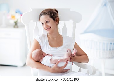 Young Mother Holding Her Newborn Child. Mom Nursing Baby. Woman And New Born Boy Relax In A White Bedroom With Rocking Chair And Blue Crib. Nursery Interior. Mother Breast Feeding Baby. Family At Home