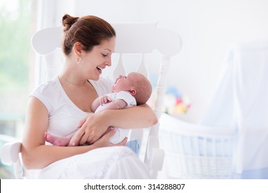 Young Mother Holding Her Newborn Child. Mom Nursing Baby. Woman And New Born Boy Relax In A White Bedroom With Rocking Chair And Blue Crib. Nursery Interior. Mother Breast Feeding Baby. Family At Home