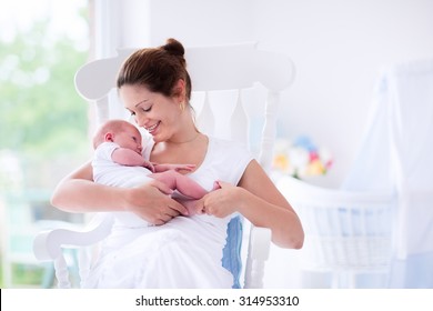 Young Mother Holding Her Newborn Child. Mom Nursing Baby. Woman And New Born Boy Relax In A White Bedroom With Rocking Chair And Blue Crib. Nursery Interior. Mother Breast Feeding Baby. Family At Home