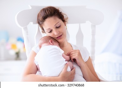 Young Mother Holding Her Newborn Child. Mom Nursing Baby. Woman And New Born Boy Relax In A White Bedroom With Rocking Chair And Blue Crib. Nursery Interior. Mother Breast Feeding Baby. Family At Home