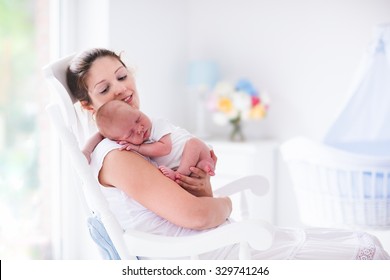 Young Mother Holding Her Newborn Child. Mom Nursing Baby. Woman And New Born Boy Relax In A White Bedroom With Rocking Chair And Blue Crib. Nursery Interior. Mother Breast Feeding Baby. Family At Home