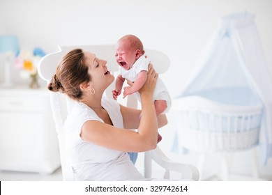 Young mother holding her newborn child. Mother comforting crying hungry baby. Woman and new born boy relax in a white bedroom with rocking chair and blue crib. Nursery interior. Family at home.