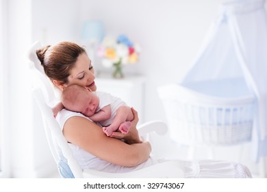 Young Mother Holding Her Newborn Child. Mom Nursing Baby. Woman And New Born Boy Relax In A White Bedroom With Rocking Chair And Blue Crib. Nursery Interior. Mother Breast Feeding Baby. Family At Home