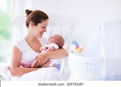 Young Mother Holding Her Newborn Child. Mom Nursing Baby. Woman And New Born Boy Relax In A White Bedroom With Rocking Chair And Blue Crib. Nursery Interior. Mother Breast Feeding Baby. Family At Home