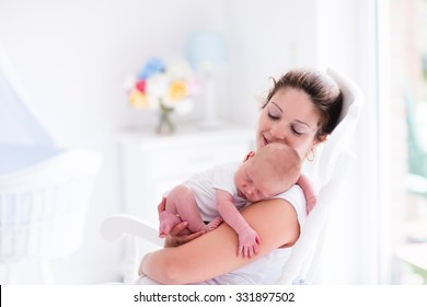 Young Mother Holding Her Newborn Child. Mom Nursing Baby. Woman And New Born Boy Relax In A White Bedroom With Rocking Chair And Blue Crib. Nursery Interior. Mother Breast Feeding Baby. Family At Home