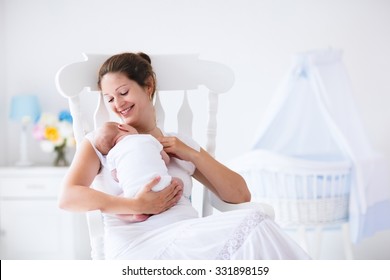 Young Mother Holding Her Newborn Child. Mom Nursing Baby. Woman And New Born Boy Relax In A White Bedroom With Rocking Chair And Blue Crib. Nursery Interior. Mother Breast Feeding Baby. Family At Home