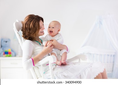 Young Mother Holding Her Newborn Child. Mom Nursing Baby. Woman And New Born Boy In White Bedroom With Rocking Chair And Blue Crib. Nursery Interior. Mother Playing With Laughing Kid. Family At Home