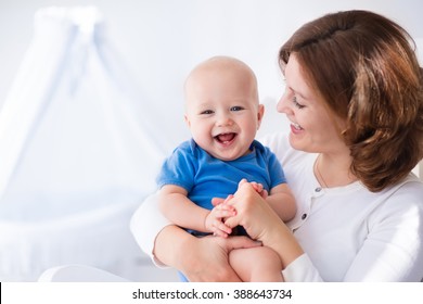 Young Mother Holding Her Newborn Child. Mom Nursing Baby. Woman And New Born Boy In White Bedroom With Rocking Chair And Blue Crib. Nursery Interior. Mother Playing With Laughing Kid. Family At Home